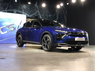 De strakke blauwe Citroën C5 X staat centraal in een overdekte showroom, verlicht door de gloed van een groot scherm op de achtergrond.