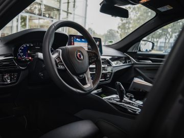 Binnenaanzicht van een moderne BMW Alpina met een stuur, een digitaal dashboard, een infotainmentsysteem en verschillende bedieningselementen. De ramen op de achtergrond zorgen voor natuurlijk licht en benadrukken het strakke design.