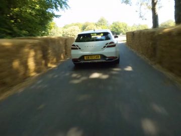 Een Genesis G70 Shooting Brake rijdt op een zonnige dag tussen hooibalen op een smalle weg omgeven door bomen.