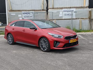 Een rode Kia Proceed-stationwagen staat geparkeerd op een verhard terrein voor gestapelde bouwmaterialen en een zwarte muur.