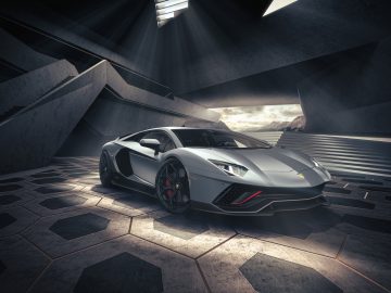 Een zilveren Lamborghini-sportwagen met strakke, hoekige ontwerpen staat geparkeerd in een futuristische, slecht verlichte garage met geometrische patronen op de vloer en muren. Een dakraam erboven straalt licht op de auto.