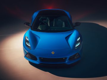 Een blauwe Lotus Emira tweedeurs sportwagen wordt verlicht in een slecht verlichte ruimte, waardoor het strakke frontontwerp en de koplampen goed tot hun recht komen.
