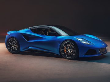 Een strakke blauwe Lotus Emira met een laag profiel, zwart dak en opvallend aerodynamisch ontwerp staat geparkeerd in een slecht verlichte omgeving.