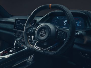 Close-up van een auto-interieur met een stuurwiel van het merk Lotus Emira, digitale displays op het dashboard en een versnellingspook op de middenconsole.