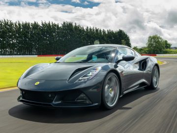 Een strakke, zwarte Lotus Emira-sportwagen rijdt op een racecircuit met groene bomen en een bewolkte lucht op de achtergrond.