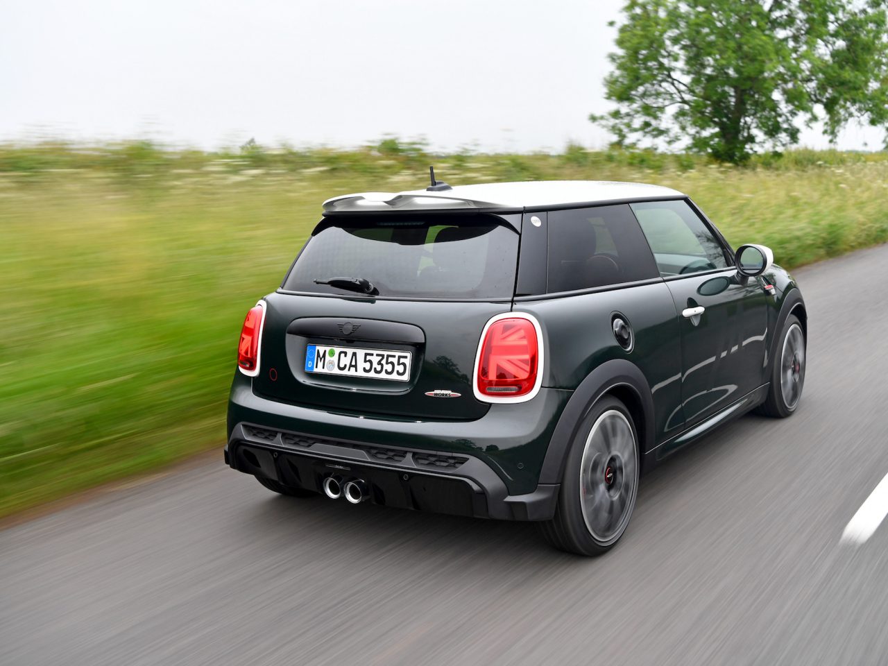 Een donkergroene MINI Anniversary Edition rijdt over een landelijke weg met groen en bomen op de achtergrond. De achterkant van de auto is zichtbaar, inclusief de kentekenplaat en de achterlichten.