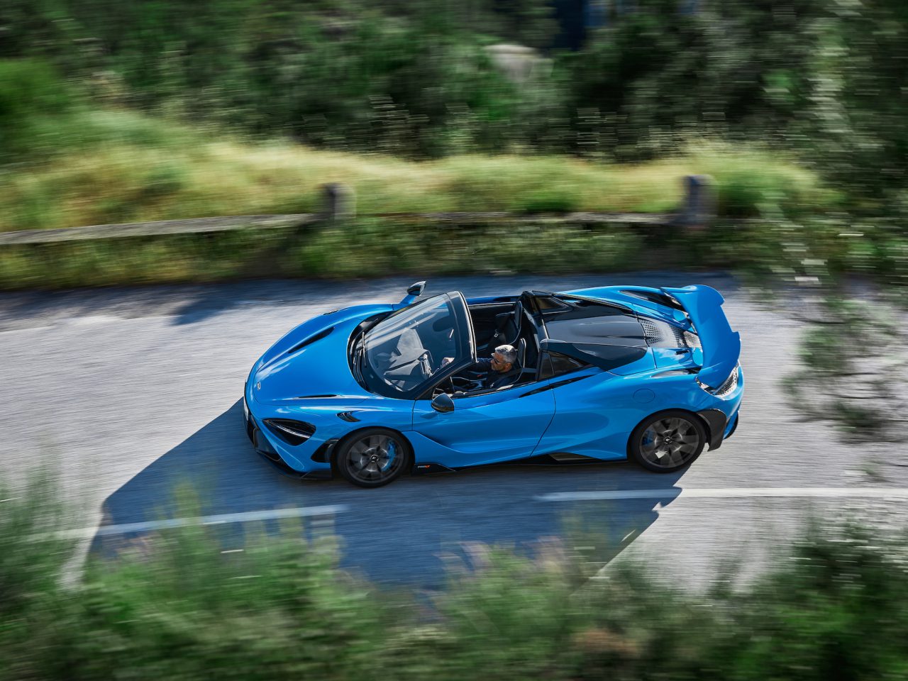 Een McLaren 765 LT Spider rijdt op een bochtige weg omgeven door groen, waarbij de bestuurder zichtbaar is door het open dak.
