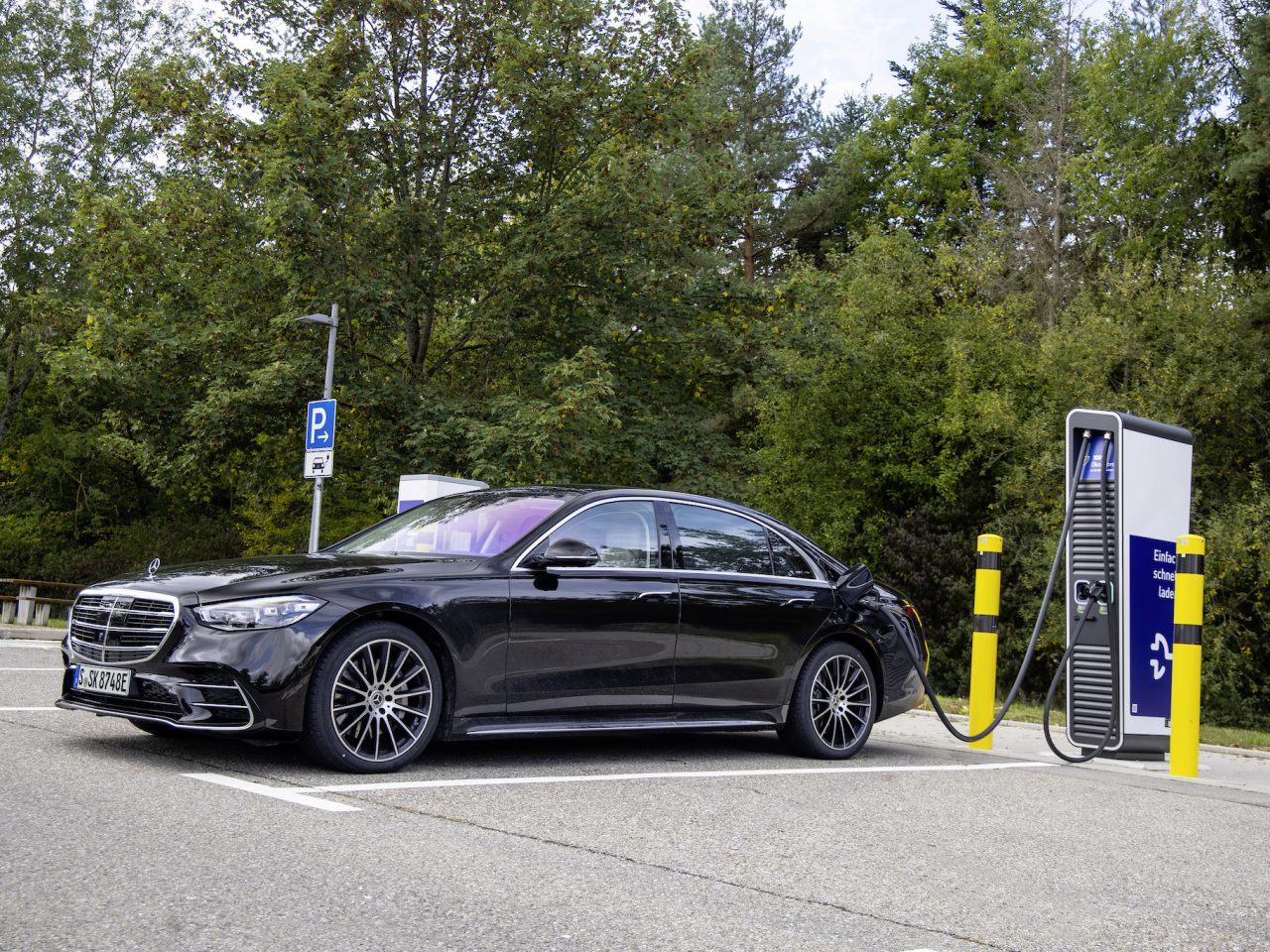Een zwarte Mercedes-Benz S 580 e staat geparkeerd en wordt opgeladen bij een laadstation voor elektrische voertuigen omgeven door bomen.
