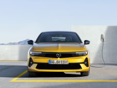 Een gele Opel Astra-auto staat met zijn gezicht naar voren op een parkeerplaats geparkeerd, met een oplaadkabel in de zijkant gestoken. Op de achtergrond is een bergachtig landschap zichtbaar.