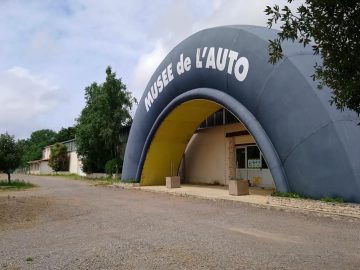 De afbeelding toont de ingang van het Musée Automobile de Vendée, een gebouw met een grote, boogvormige structuur en de naam bovenaan geschreven. De omgeving bestaat uit bomen en een grindpad.