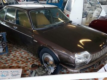 Een klassieke donkerbruine auto wordt tentoongesteld in een showroom in het Musée Automobile de Vendée. De auto heeft een retro-ontwerp met een vierdeurs carrosserie. Rondom de auto bevinden zich andere vintage voertuigen en mechanische componenten, waardoor een sfeer van tijdloze autogeschiedenis ontstaat.