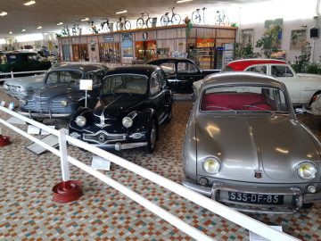 Een tentoonstelling van oldtimers siert de showroom met een tegelvloer, terwijl fietsen op de achtergrond aan bagagerekken hangen, waardoor een sfeer ontstaat die doet denken aan Musée Automobile de Vendée.