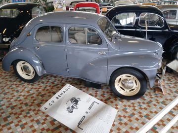 In het Musée Automobile de Vendée staat een lichtblauwe Renault 4CV, omringd door andere vintage voertuigen. Informatie over de auto staat op een bord ervoor.