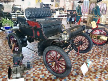 Een vintage zwarte auto met rode wielen wordt tentoongesteld in het Musée Automobile de Vendée. De auto heeft een getuft lederen stoel en decoratieve accenten. Op de achtergrond zijn andere vintage items en vitrines zichtbaar.