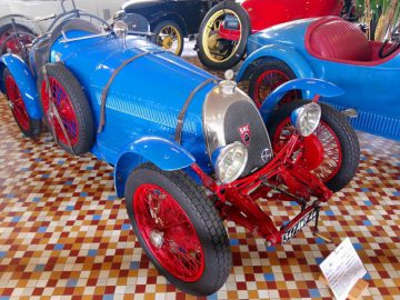 Een blauwe vintage raceauto met rode wielen en accenten wordt binnenshuis tentoongesteld in het Musée Automobile de Vendée. De auto heeft een klassiek, afgerond ontwerp en is voorzien van een kentekenplaat aan de voorzijde. Op de achtergrond zijn andere auto's zichtbaar.