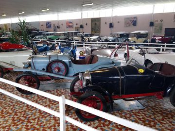 Een verzameling oldtimers die binnenshuis worden tentoongesteld op een tegelvloer met witte balustrades die ze scheiden, in de goed verlichte omgeving van het Musée Automobile de Vendée.