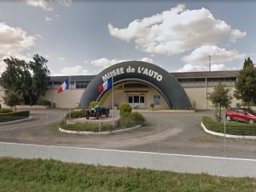 Buitenaanzicht van het Musée Automobile de Vendée met Franse en EU-vlaggen, een boogvormige ingang en buiten een oldtimer.