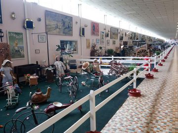 Een kamer vol antieke voorwerpen, waaronder fietsen, mannequins en diverse verzamelobjecten uit het Musée Automobile de Vendée, tentoongesteld achter een wit hek. De ruimte is goed verlicht met veel ingelijste foto's aan de muren.