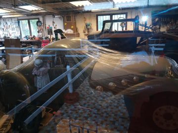 Een werkplaats met twee oldtimers, waarvan één bedekt met plastic. Op de achtergrond hangen verschillende gereedschappen aan de muur en werkbanken met apparatuur, die doen denken aan de gedetailleerde displays in Musée Automobile de Vendée.