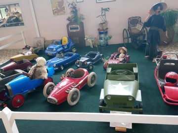 Een collectie vintage kinderautootjes is te zien in een overdekte tentoonstelling in het Musée Automobile de Vendée, met verschillende stijlen en kleuren. Op de achtergrond zijn ook een volwassen mannequin en kinderwagen zichtbaar.