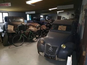 Een slecht verlichte kamer in het Musée Automobile de Vendée toont verschillende oldtimers, fietsen en andere antieke voertuigen. Op de linkermuur is een bord "INFIRMERIE" zichtbaar. Het tafereel ademt een oude, nostalgische sfeer.