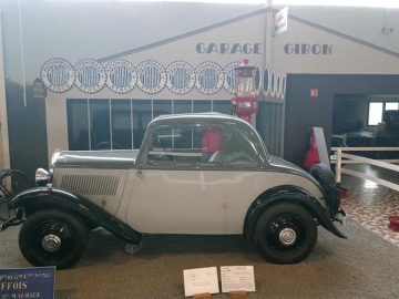 In het Musée Automobile de Vendée is een vintage grijze auto te zien, compleet met een retro garageachtergrond, een ouderwetse benzinepomp en een paar informatieborden voor de auto.