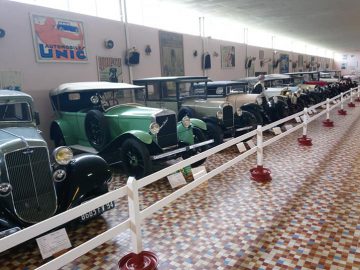 Een rij oldtimers wordt tentoongesteld in een museumomgeving met een tegelvloer in het Musée Automobile de Vendée. De auto's staan opgesteld langs een slagboom met informatieborden ervoor. Vintage posters sieren de muur erachter.