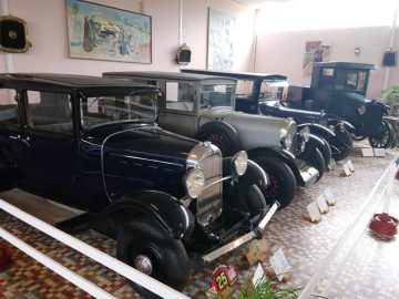 In het Musée Automobile de Vendée staat een rij oldtimers tentoongesteld in een museum, gescheiden door witte balustrades. De vloer is betegeld en de muren zijn voorzien van kunstwerken en vintage items. Voor elke auto worden tags met informatie geplaatst.