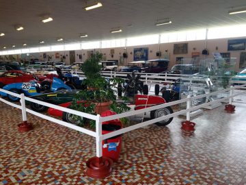 Een binnententoonstelling in het Musée Automobile de Vendée toont verschillende klassieke auto's en motorfietsen, omgeven door witte balustrades en planten op een tegelvloer.