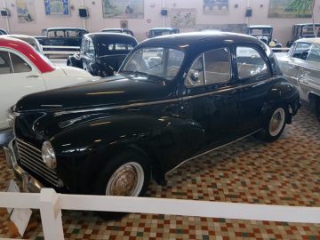 Een zwarte vintage auto wordt binnen tentoongesteld tussen andere klassieke auto's op een vloer met patronen in het Musée Automobile de Vendée. Voor de auto is een bord zichtbaar.