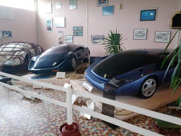 Twee futuristische, aerodynamische auto's worden tentoongesteld in een kamer met ingelijste foto's en planten in het Musée Automobile de Vendée. Op de voorgrond zijn een wit hekwerk en een geblokte vloer zichtbaar, wat bijdraagt aan de unieke charme van het museum.