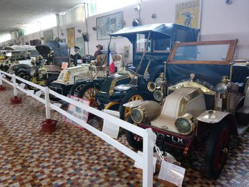 In het overdekte Musée Automobile de Vendée staat een rij oldtimers tentoongesteld, achter een witte balustrade op een tegelvloer. Elke auto heeft beschrijvende tags en posters op de muur erachter.