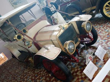 Een vintage witte en rode auto met koperen accenten, tentoongesteld in het Musée Automobile de Vendée, geparkeerd op een geblokte vloer. Voor het voertuig zijn informatieborden geplaatst.