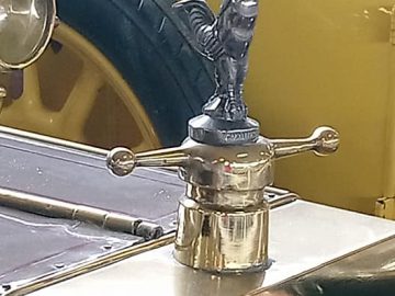 Close-up van een vintage gele auto in het Musée Automobile de Vendée, met een decoratief ornament op de motorkap van een haan en een koperen radiatordop, met een stuur en leren stoelen op de achtergrond.