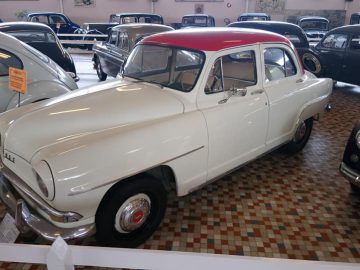 Een vintage witte en rode auto wordt tentoongesteld in een overdekte showroom in het Musée Automobile de Vendée, omringd door andere klassieke auto's op een tegelvloer met patroon.