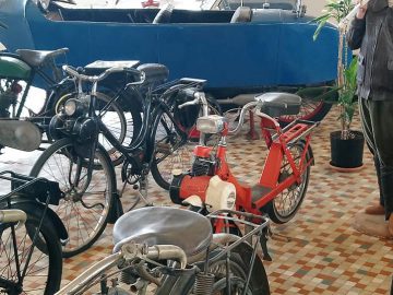 Een verzameling vintage motorfietsen en een blauwe klassieke auto, binnenshuis tentoongesteld op een tegelvloer in het Musée Automobile de Vendée, met vlakbij een mannequin gekleed in een leren jas en pet.