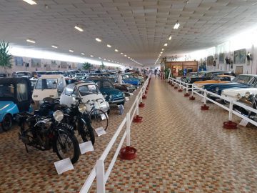 Het Musée Automobile de Vendée beschikt over een grote overdekte tentoonstellingsruimte met rijen oldtimers en motorfietsen op een tegelvloer, gescheiden door witte balustrades.