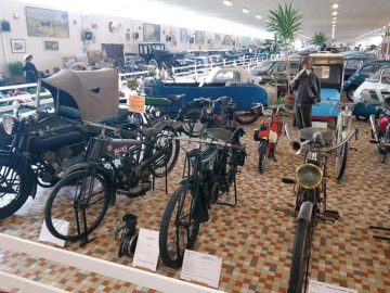 Een ruime overdekte tentoonstelling in het Musée Automobile de Vendée toont verschillende vintage motorfietsen en een paar klassieke auto's, allemaal in rijen gerangschikt op een tegelvloer met patroon en informatieve borden.