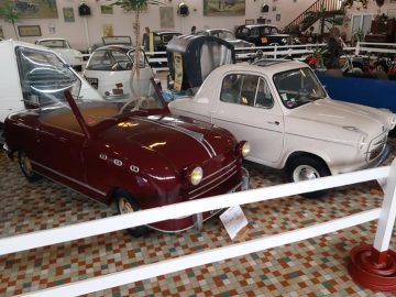 Een verzameling vintage brommobielen, waaronder een rood en een wit model, wordt prachtig tentoongesteld in het Musée Automobile de Vendée, met tegelvloeren en diverse andere klassieke voertuigen op de achtergrond.