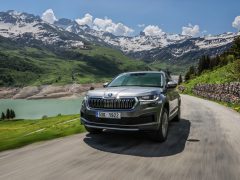 Een grijze Skoda Kodiaq rijdt over een weg door een bergachtig landschap met een meer en met sneeuw bedekte toppen op de achtergrond.