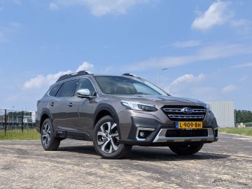 Een grijze Subaru Outback staat geparkeerd op een vlak, open terrein, omlijst door een heldere hemel met een paar wolken die lui op de achtergrond drijven. De auto pronkt trots met een geel kenteken, versierd met zwarte letters en cijfers, beginnend met ‘L-909-BH.