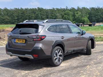 Een grijze Subaru Outback staat geparkeerd op een verhard oppervlak met een groene bosachtergrond. De Subaru Outback is voorzien van een zichtbaar kenteken "L-909-BH.