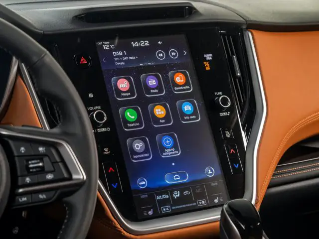 Close-up van het infotainmentsysteem van een Subaru Outback met verschillende apps op een touchscreen. Het dashboard is voorzien van een mix van zwarte en lichtbruine materialen, met bedieningsknoppen die het scherm flankeren. Stuur in zicht.