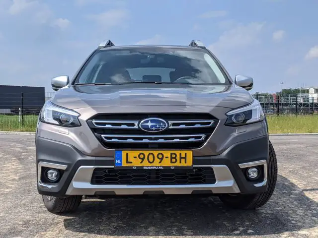 Een vooraanzicht van een grijze Subaru Outback SUV met een kentekenplaat met de tekst "L-909-BH", geparkeerd op een asfaltoppervlak tegen een achtergrond van een grasveld en gebouwen.