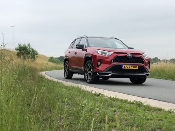 Een rode Toyota RAV4 Plug-in Hybrid SUV staat op een bewolkte dag aan de kant van een verharde weg geparkeerd, met gras en struiken langs de weg. Het voertuig heeft een Europees kenteken.