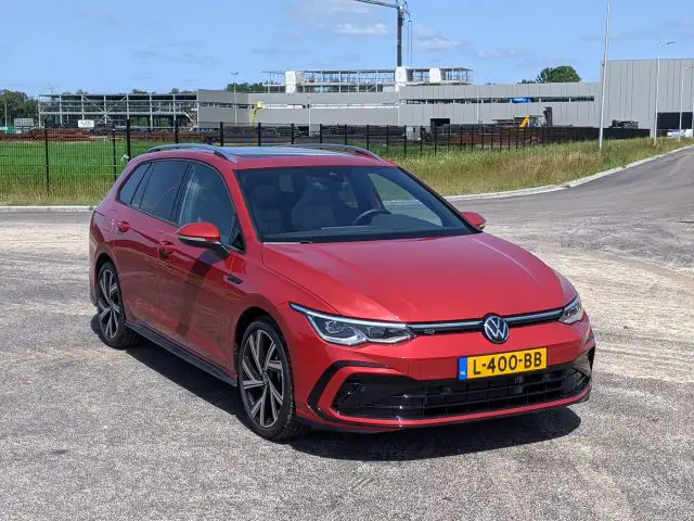 Een rode Volkswagen Golf Variant stationwagen staat geparkeerd op een verhard terrein met industriële gebouwen en een gedeeltelijk bewolkte lucht op de achtergrond. De auto heeft een geel Europees kenteken.