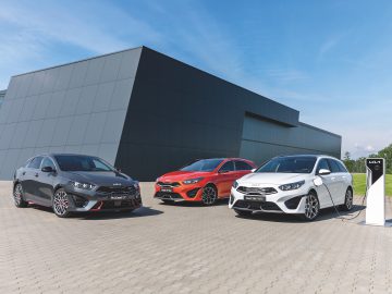 Voor een modern gebouw staan drie auto's geparkeerd, waaronder een strakke Kia Ceed; één is aangesloten op een laadstation. De auto's zijn grijs, rood en wit en het tafereel speelt zich af onder een helderblauwe lucht.