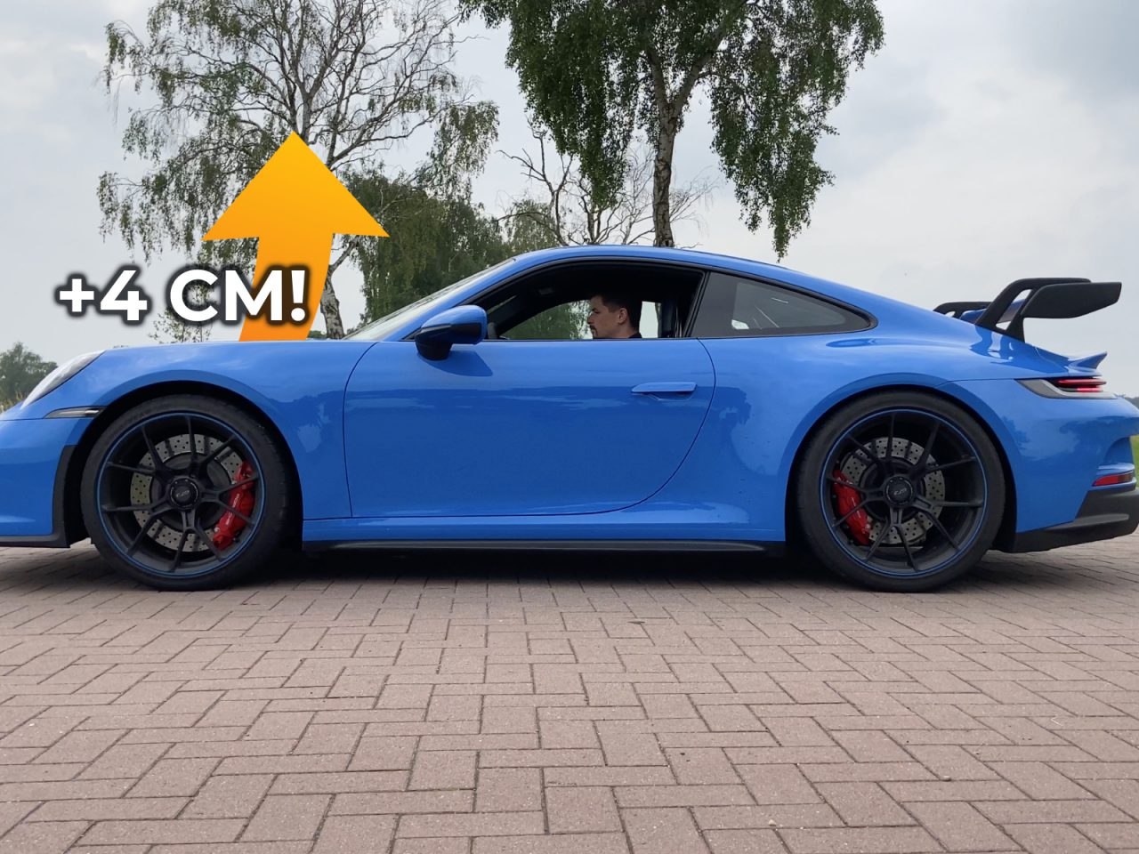 Buiten staat op een stenen ondergrond een blauwe Porsche 911 GT3 geparkeerd, waarbij een pijl een 4 cm hogere rijhoogte aangeeft. De auto is voorzien van zwarte wielen en rode remklauwen.