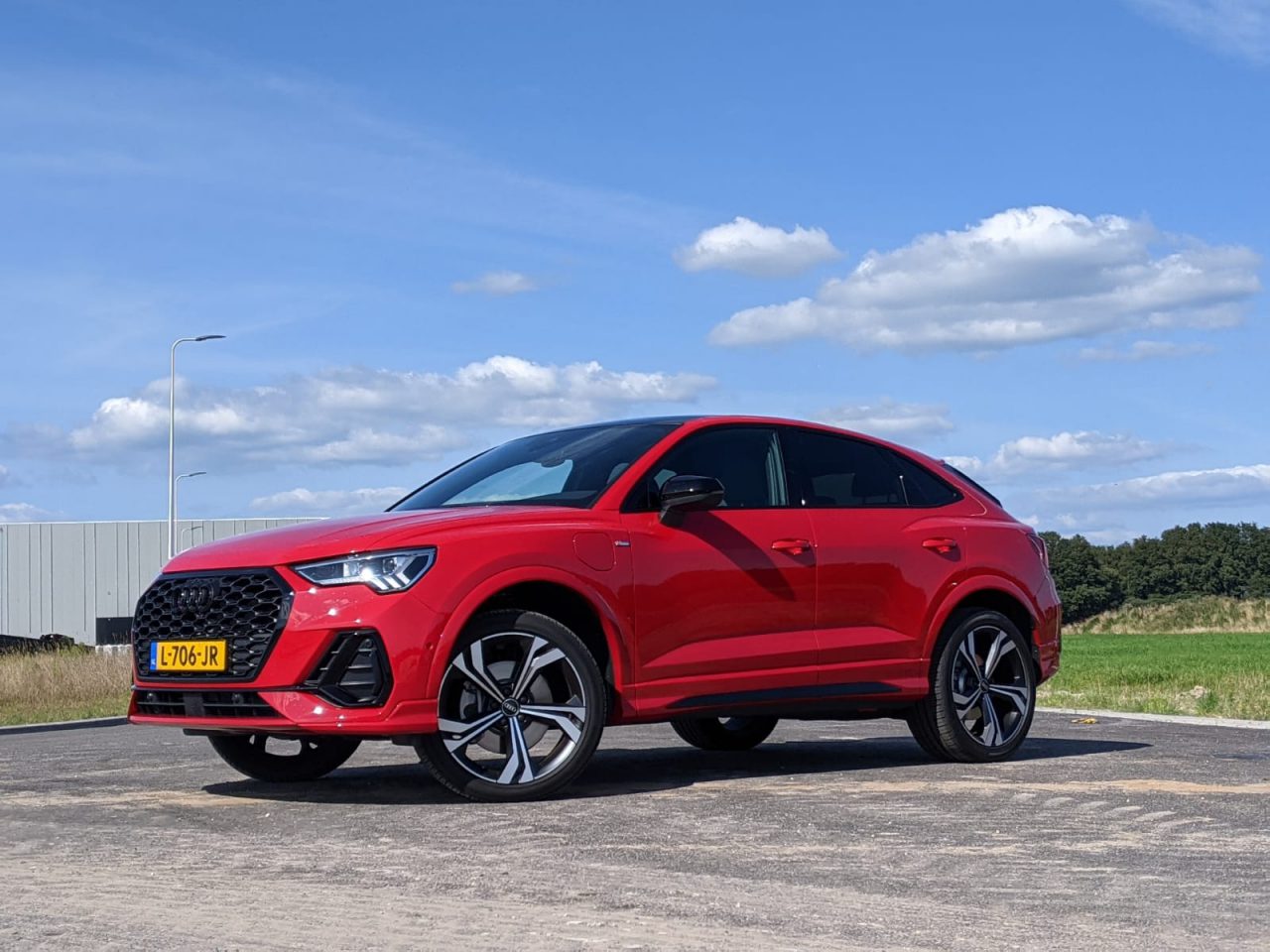 Op een verhard terrein staat een rode Audi Q3 plug-in hybride met zwarte accenten geparkeerd. Het voertuig heeft een kentekenplaat en speelt zich af tegen een achtergrond van een gedeeltelijk bewolkte blauwe lucht en groen.