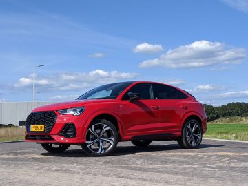 Op een verhard terrein staat een rode Audi Q3 plug-in hybride met zwarte accenten geparkeerd. Het voertuig heeft een kentekenplaat en speelt zich af tegen een achtergrond van een gedeeltelijk bewolkte blauwe lucht en groen.